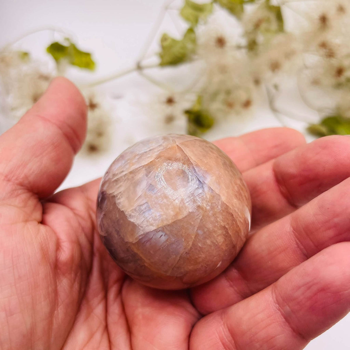 Nahaufnahme der Peach-Mondstein-Kugel in einer Hand mit schimmernden Kristallstrukturen und glänzend polierter Oberfläche.
