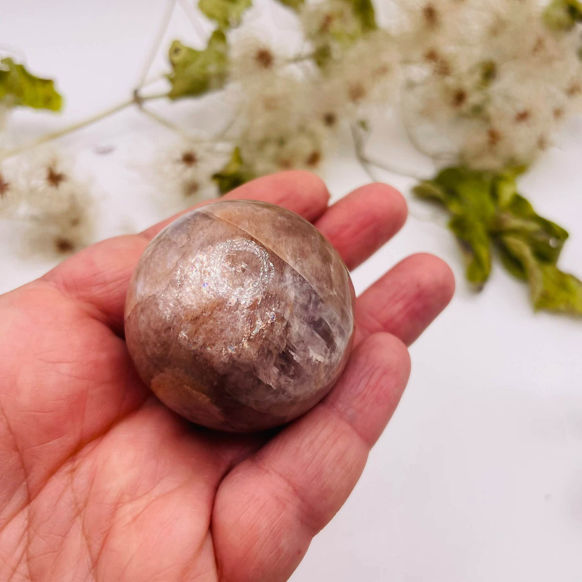 Weitere Detailansicht der Peach-Mondstein-Kugel in einer Hand mit schimmernden Kristallstrukturen und glänzend polierter Oberfläche.
